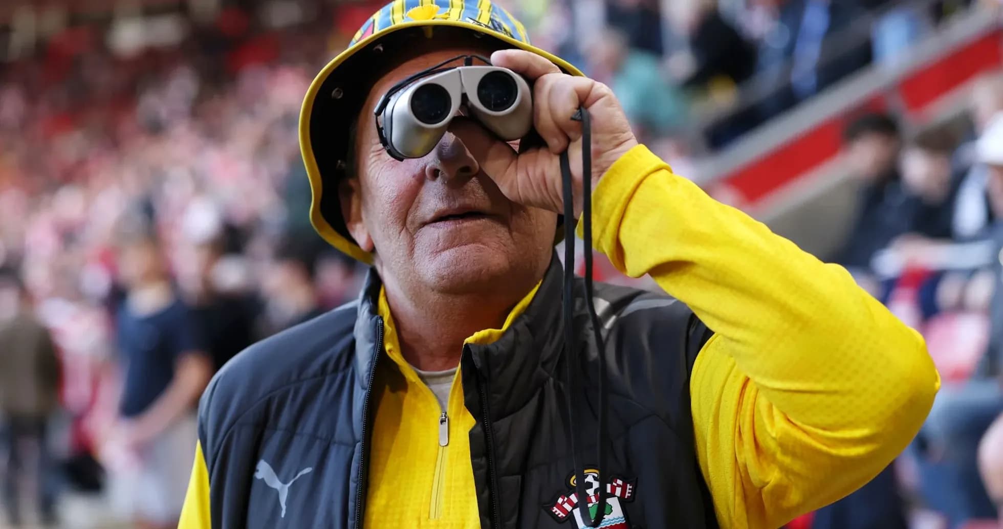 A fan wearing binoculars and a hat at a match A fan wearing binoculars and a hat at a match